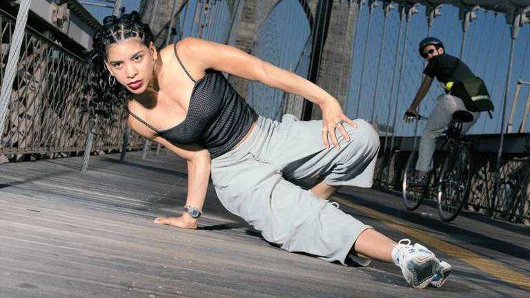 Woman dancer has one hand on the ground and one on her leg as she performs a move on the Brooklyn Bridge with bicyclist noticing.