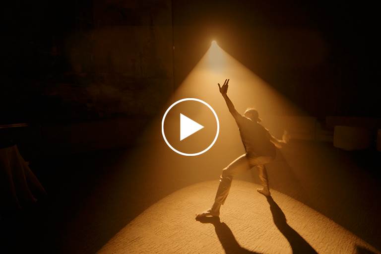 Dancer in spotlight casting a shadow performs solo. Play button is superimposed on image.