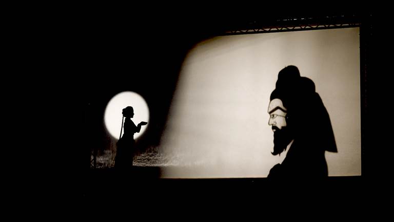 A woman stands on the left with her hands cupped. To the right is a shadow of a man鈥檚 head behind a large white screen. The man has a long beard and raised eyebrows that stretch across the side of his face. 