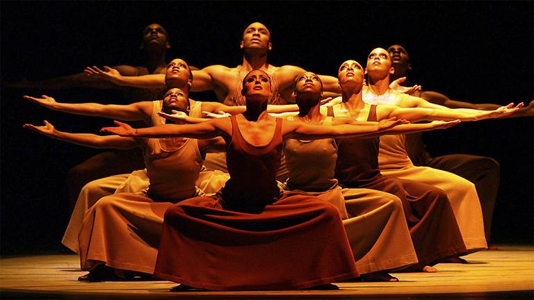 Nine Alvin Ailey dancers on stage with arms outstretched looking up into a warm glowing light.