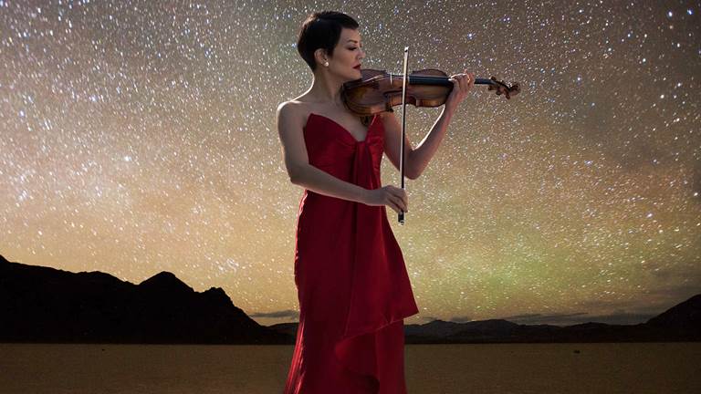 A light-skinned woman in a red strapless evening gown plays a violin in front of a starry sky over a mountain range
