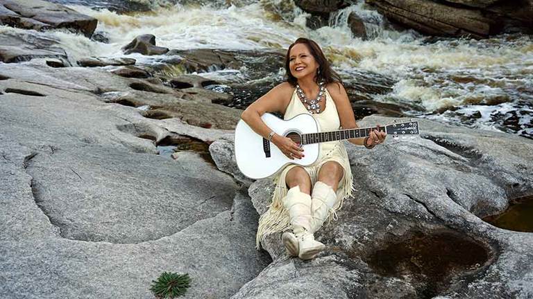 Woman in Native American dress plays a guitar while sitting on rocks in a stream.