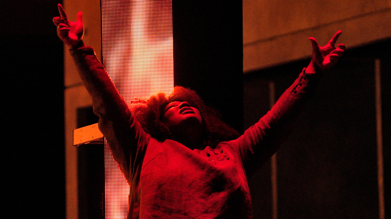 A woman in a tunic raises her arms to the sky in a wash of red light